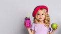 Blonde kid in red hat and purple blouse. She smiling, holding pink cocktail bottle and green apple, posing isolated on white Royalty Free Stock Photo