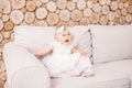 Little blue-eyed girl blond in a white tulle dress with a decoration on her head playing and rejoicing on a beige sofa in a room w Royalty Free Stock Photo