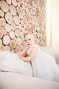 Little blue-eyed girl blond in a white tulle dress with a decoration on her head playing and rejoicing on a beige sofa in a room w Royalty Free Stock Photo