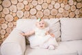 Little blue-eyed girl blond in a white tulle dress with a decoration on her head playing and rejoicing on a beige sofa in a room w Royalty Free Stock Photo