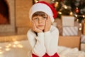 Little bored girl waiting for Christmas, sitting in festive living room, keeping hands on cheek, looks at camera with sad