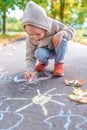 Little boy in autumn on street, draws on pavement, multicolored crayons. Warm clothes on a cold day, beige cardigan with