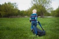 The little boy with blond hair pulling a large backpack chery on the green grass in nature, travel, baby, adventure Royalty Free Stock Photo