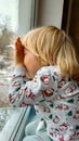 A little boy with blonde hair sits by the window on the windowsill and looks out the window. Winter. New Year& x27;s Royalty Free Stock Photo