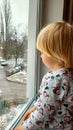 A little boy with blonde hair sits by the window on the windowsill and looks out the window. Winter. New Year& x27;s Royalty Free Stock Photo