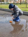 A little boy in blue rubber boots launches an orange paper boat in a puddle. The boy is having fun and smiling Royalty Free Stock Photo