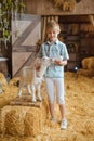 A little boy in a blue shirt and shorts stands in front of the wooden doors of a barn and feeds a white baby goat Royalty Free Stock Photo