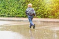 Little boy child in raincoat,rubber boots running playing in puddle in rainy summer park.Kid jumping outdoor by autumn