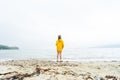 A little boy child in a yellow windbreaker stands on the sandy shore of the ocean all alone.