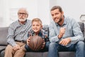 Little boy on couch with grandfather and father, cheering for a basketball game and holding a Royalty Free Stock Photo
