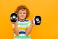 Little boy with curly hair in colourful t-shirt and shorts holds small music columns and dances isolated on yellow