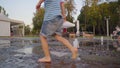 A little boy in denim shorts and yellow shoes runs in the fountain on summer evening