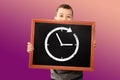 Little boy of European appearance, a schoolboy, with two hands holds a black school board with a clock and an arrow on a light Royalty Free Stock Photo