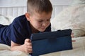 little boy at expressive face using a digital tablet in bed plays games, watch cartoons, talks with friends, writes a
