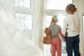 Little boy and girl in home clothes holding hands, looking out of a large window while spending time together at home Royalty Free Stock Photo