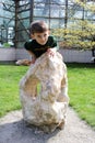 A little boy in a green sweater with dinosaurs is trying to climb a large stone, which is located near a glass building
