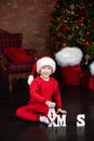 Little boy in hat red Santa sit on floor by Christmas tree and playing wooden letters Xmas. Happy small boy decorated festive livi Royalty Free Stock Photo