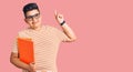 Little boy kid holding book wearing glasses with a big smile on face, pointing with hand finger to the side looking at the camera Royalty Free Stock Photo