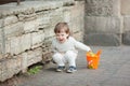 Little boy with long blond hair crying standing on the street. In his hand he is holding an orange bucket to play in the sandbox.