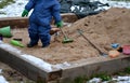 Little boy playing in blue winter overalls and green fingerless gloves on the sandpit. He holds habits and digs sand. There is a w Royalty Free Stock Photo