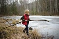 Little boy playing large branch on shore of forest lake on early spring day. Surface of lake is still under ice. Child tries to Royalty Free Stock Photo