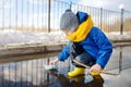 Little boy playing with origami paper boat in large puddle near town houses. Paper ship floating in water