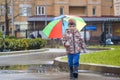 Little boy playing in rainy summer park. Child with colorful rainbow umbrella, waterproof coat and boots jumping in puddle and mud