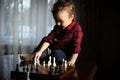 Little boy plays chess alone on the table in front of the window. Royalty Free Stock Photo