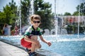 Little boy plays in the square near pool with water jets in the fountain at sunny summer day. Active summer leisure for kids in Royalty Free Stock Photo