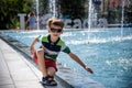 Little boy plays in the square near pool with water jets in the fountain at sunny summer day. Active summer leisure for kids in Royalty Free Stock Photo
