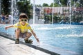 Little boy plays in the square near pool with water jets in the fountain at sunny summer day. Active summer leisure for kids in Royalty Free Stock Photo