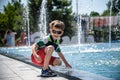 Little boy plays in the square near pool with water jets in the fountain at sunny summer day. Active summer leisure for kids in Royalty Free Stock Photo