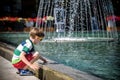 Little boy plays in the square near pool with water jets in the fountain at sunny summer day. Active summer leisure for kids in Royalty Free Stock Photo