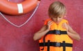 Little boy putting on orange life vest near red wall with safety ring