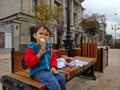 A little boy in a red-blue sweatshirt sits on a bench and eats ice cream on a streetin the city