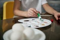 Little boy`s hand squeezing green paint from a tube onto a palette, blurry eggs on a saucer in the foreground. Closeup Royalty Free Stock Photo