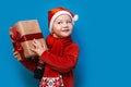 Little boy in Santa`s hat with cozy gift box and red tape want to know what inside the box, put to his ear