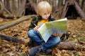 Little boy scout is orienteering in forest. Child is sitting on fallen tree and looking on map on background of teepee hut Royalty Free Stock Photo