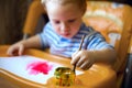 A little boy is sitting at the kids table, holding a brush, paint, paints