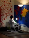 A little boy, sitting in a rocking chair, reads, and a lamp shines above him. Walls - a map of the world and an orange fun tree Royalty Free Stock Photo