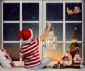 Little boy sitting on the window and looking at Santa Claus flying in his sleigh against moon sky. Royalty Free Stock Photo