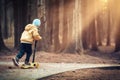 Little boy skates on scooter in evening park at sunset under light of lanterns. kid is riding scooter along path in dark forest.