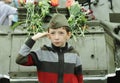 Little boy in a Soviet Army soldier side cap saluting in front of the armour of tank and flowers, celebrating the Victory Day