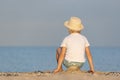 Little boy in straw hat sitting on beach and looks out to sea. Back view Royalty Free Stock Photo