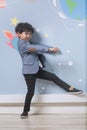 A little boy in a suit stands sideways on one leg in front of a wall