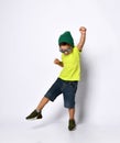 Little boy in sunglasses, green hat, yellow t-shirt, denim shorts, khaki sneakers. Raised leg up showing tongue. Isolated on white Royalty Free Stock Photo