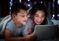 Little boy and teenage sibling sister holding digital tablet while lying under blanket in the dark at night reading