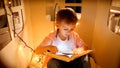 Little boy in toy house with light garlands reading book at night. Concept of child education and reading in dark room Royalty Free Stock Photo