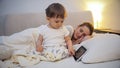 Cute little boy using digital tablet while his mother sleeping in bed Royalty Free Stock Photo