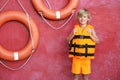 Little boy wearing orange life vest near red wall with safety rings Royalty Free Stock Photo